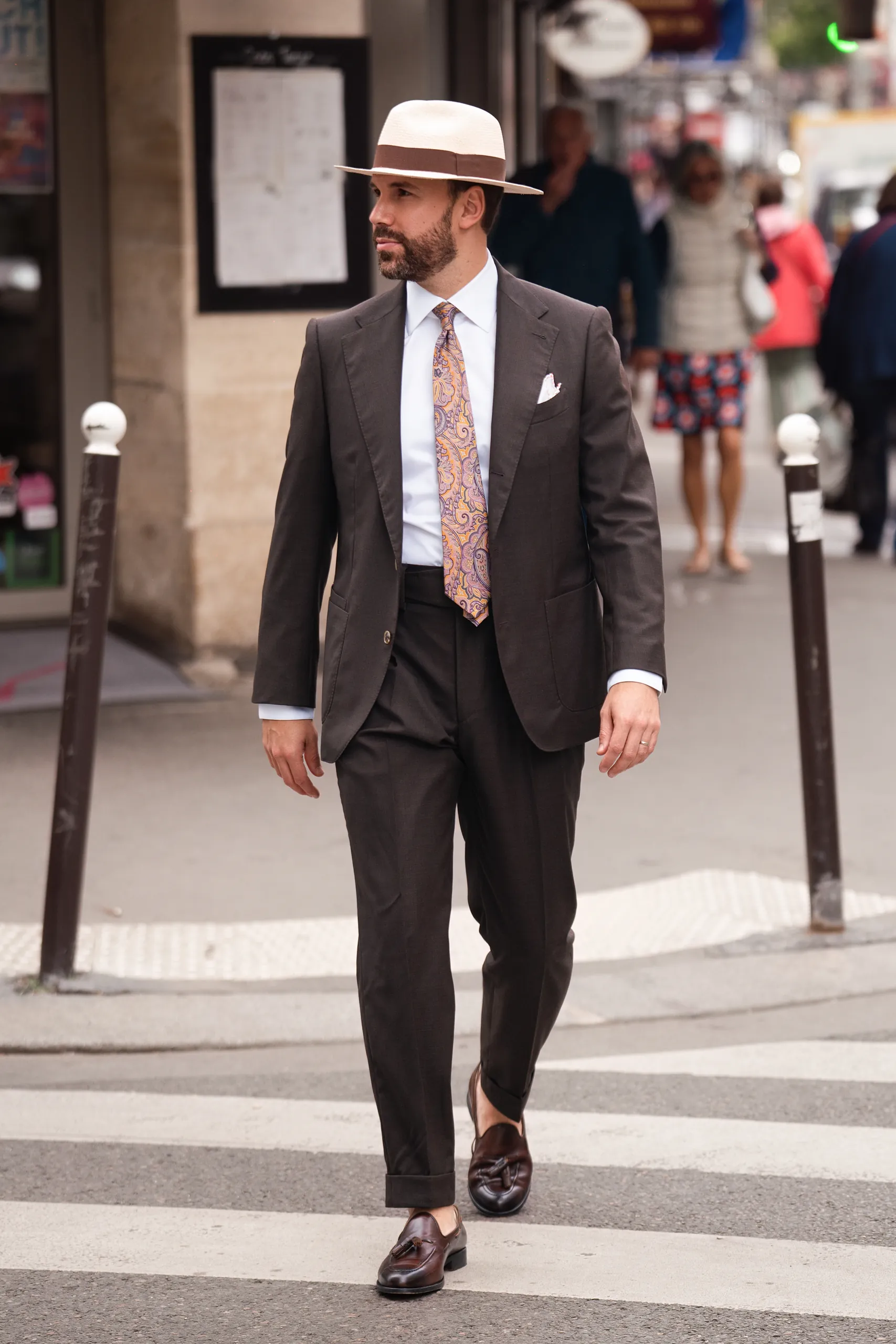 Homme en costume sur mesure brun et chapeau panama traversant une rue parisienne