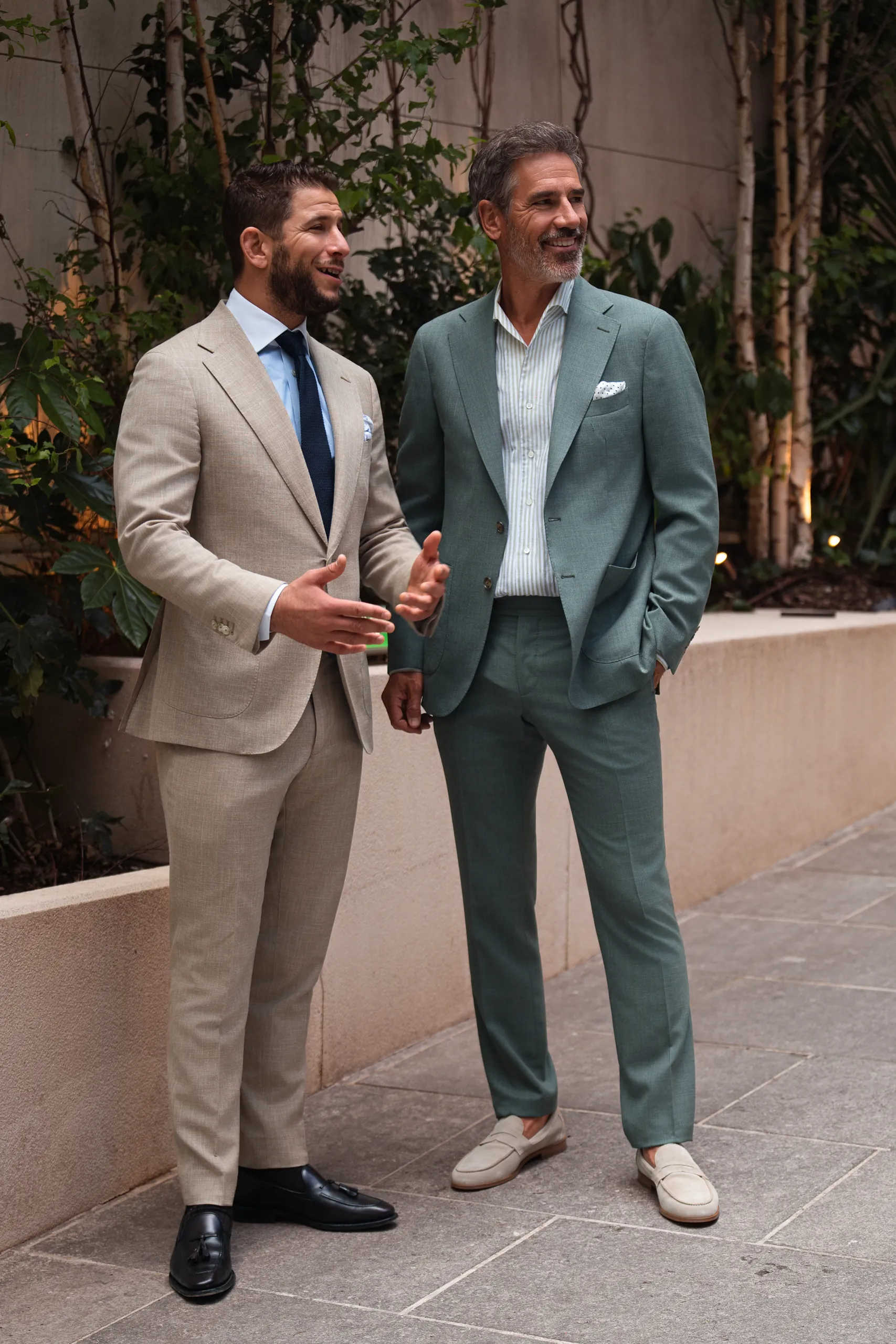 Deux hommes en costume sur mesure, l'un beige, l'autre vert sauge, sur une terrasse végétalisée