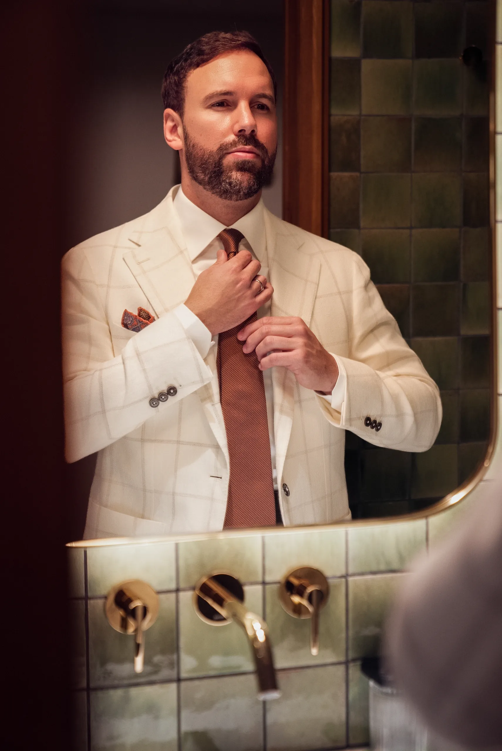 Homme ajustant son nœud de cravate devant un miroir, veste sur mesure blanche à carreaux