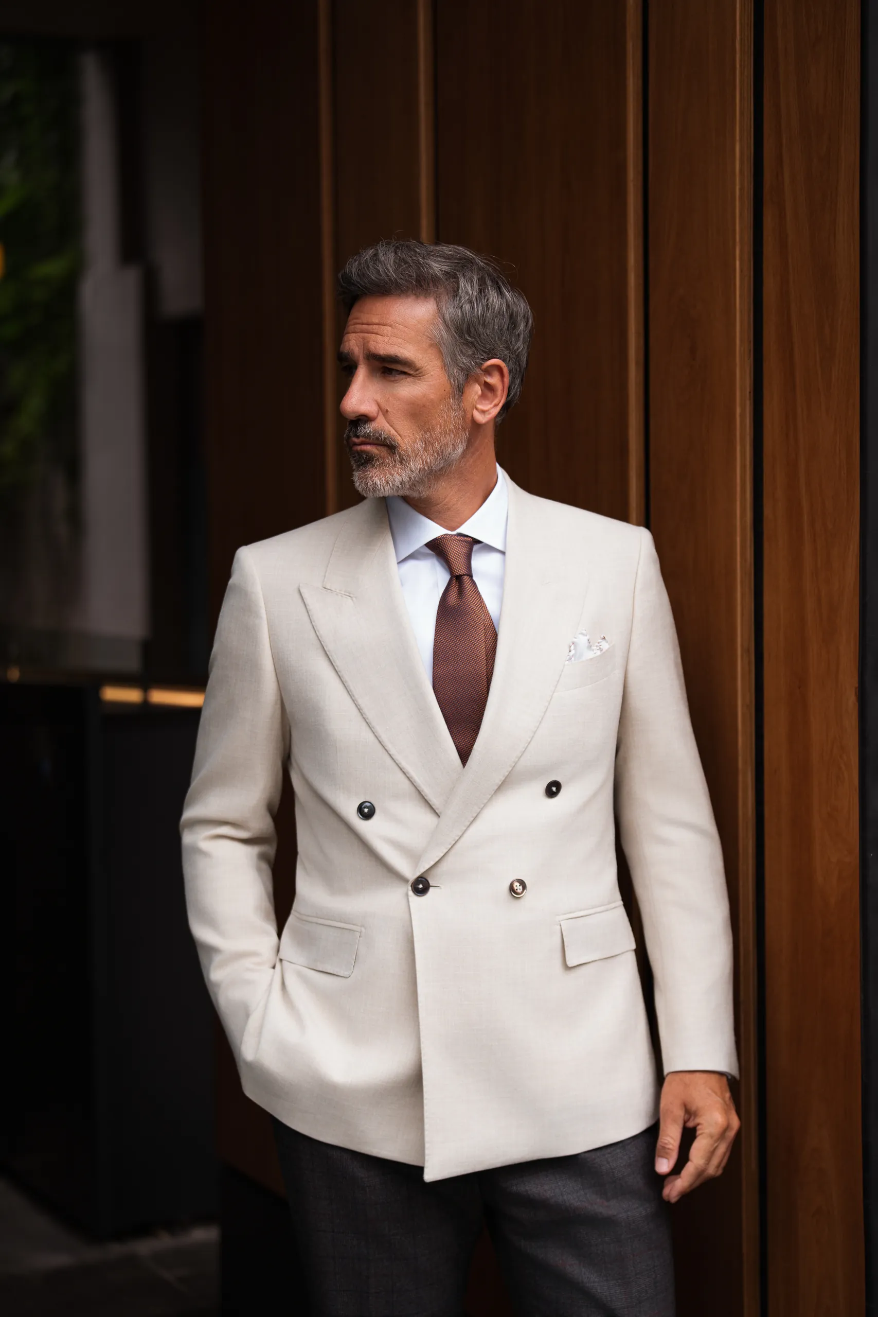 Veste croisée sur mesure ivoire et cravate brune, devant des boiseries