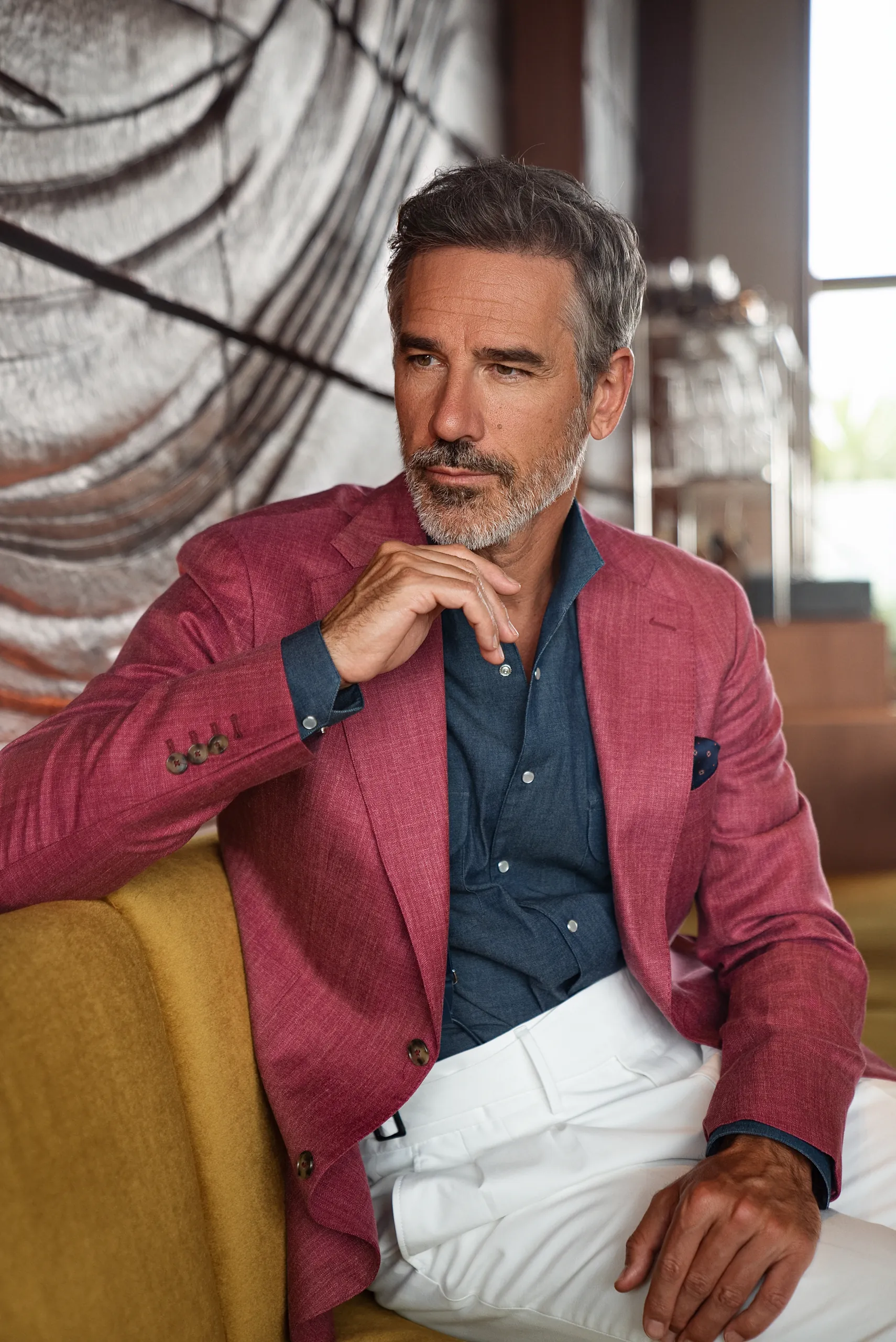 Veste sur mesure framboise, chemise en jean et pantalon blanc, assis dans un fauteuil jaune